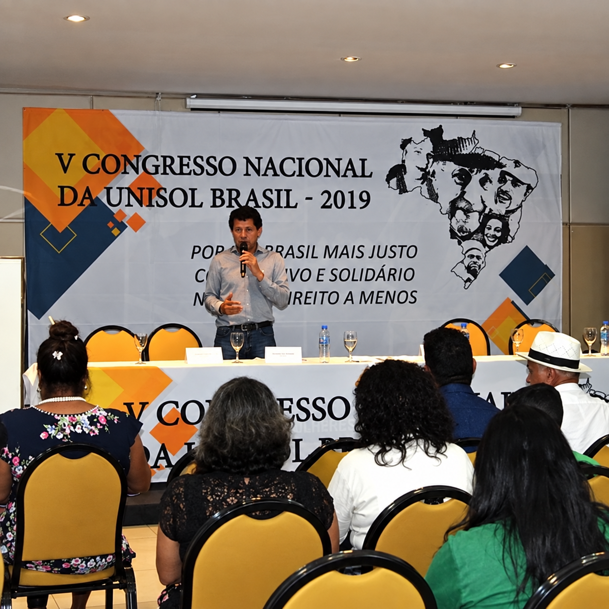 Banner backdrop personalizado em tecido para evento corporativo, palestrante ao microfone durante congresso com plateia ao fundo